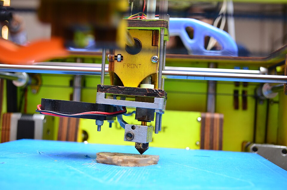 A 3D printer nozzle depositing the first layers of a small object onto a blue build plate