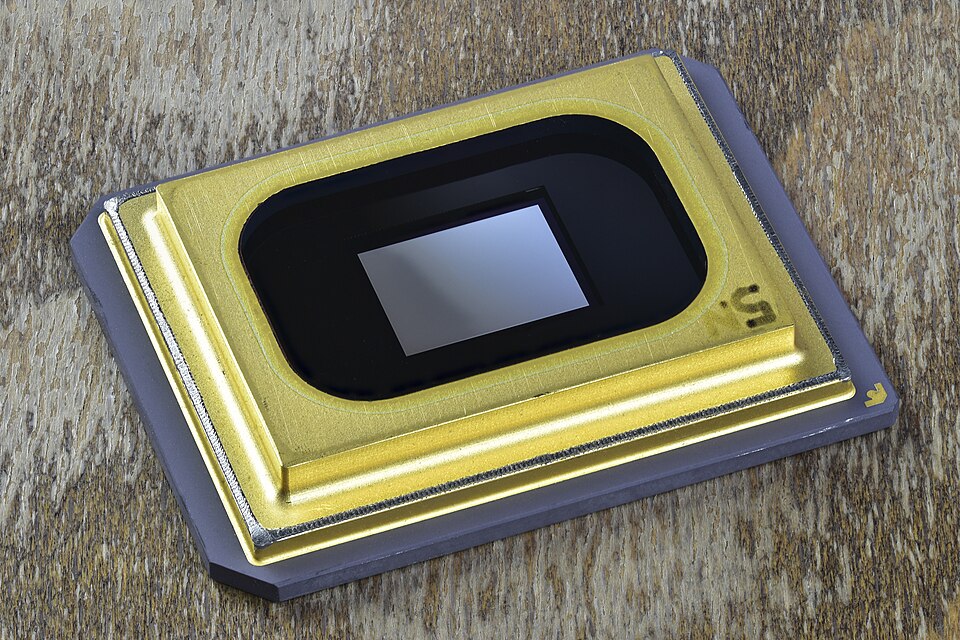 A DLP DMD chip with a gold frame around a small glass window over the mirror array