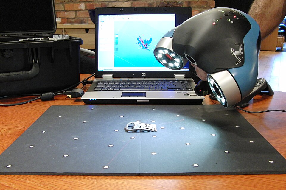 A handheld 3D scanner with two ring lights pointed at a small object on a dotted mat, with the 3D model appearing on a laptop