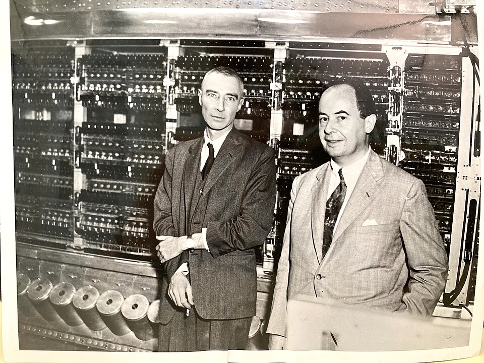 Two men in suits standing in front of a large computer made of racks of vacuum tubes