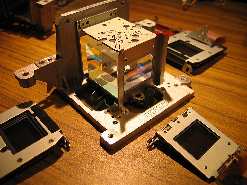 The inside of an LCD projector opened up: a glass prism cube in a metal frame, with three small LCD panels beside it