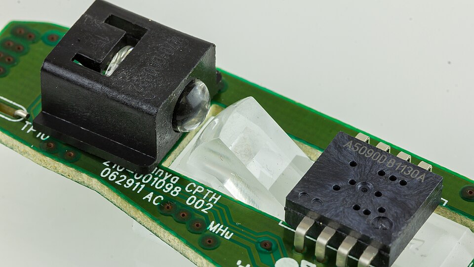 The inside of an optical mouse: a black LED housing, a clear plastic lens and a square black sensor chip on a green circuit board