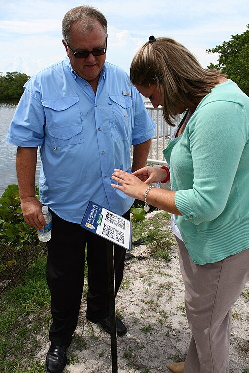 A visitor holds a smartphone over a sign on a post that carries two QR codes, while a guide watches