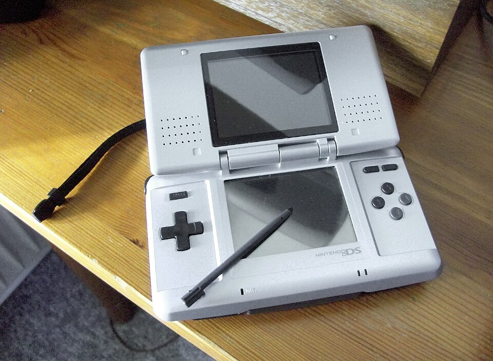 A silver handheld games console opened on a desk, with a plastic stylus resting on its lower touch screen