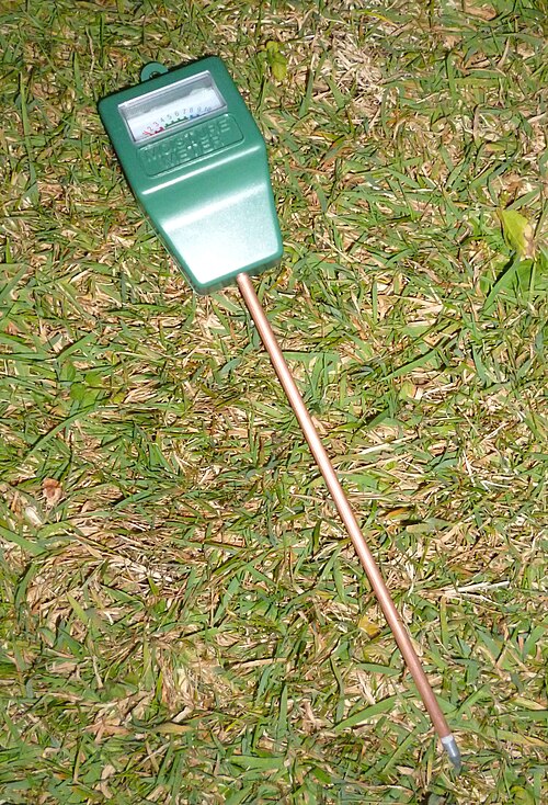 A green soil moisture meter with a metal probe pushed into grass