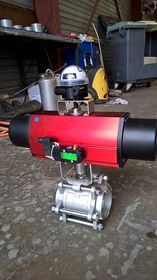 A red industrial valve actuator on a steel ball valve, with a small black solenoid valve mounted on its side