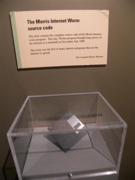 A floppy disk in a clear display case below a museum card titled The Morris Internet Worm source code