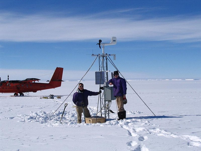 Two people beside an automatic weather station mast standing in snow, with a small aircraft behind them