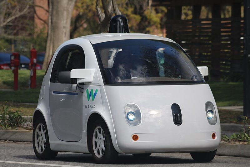 A small white two-seat driverless car with a sensor dome on its roof, driving on a street