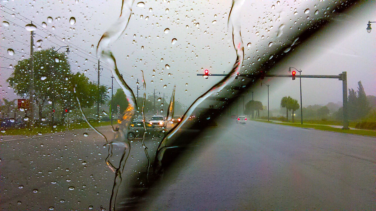 A rain-covered car windscreen with a clear arc just swept by the wiper, traffic ahead.