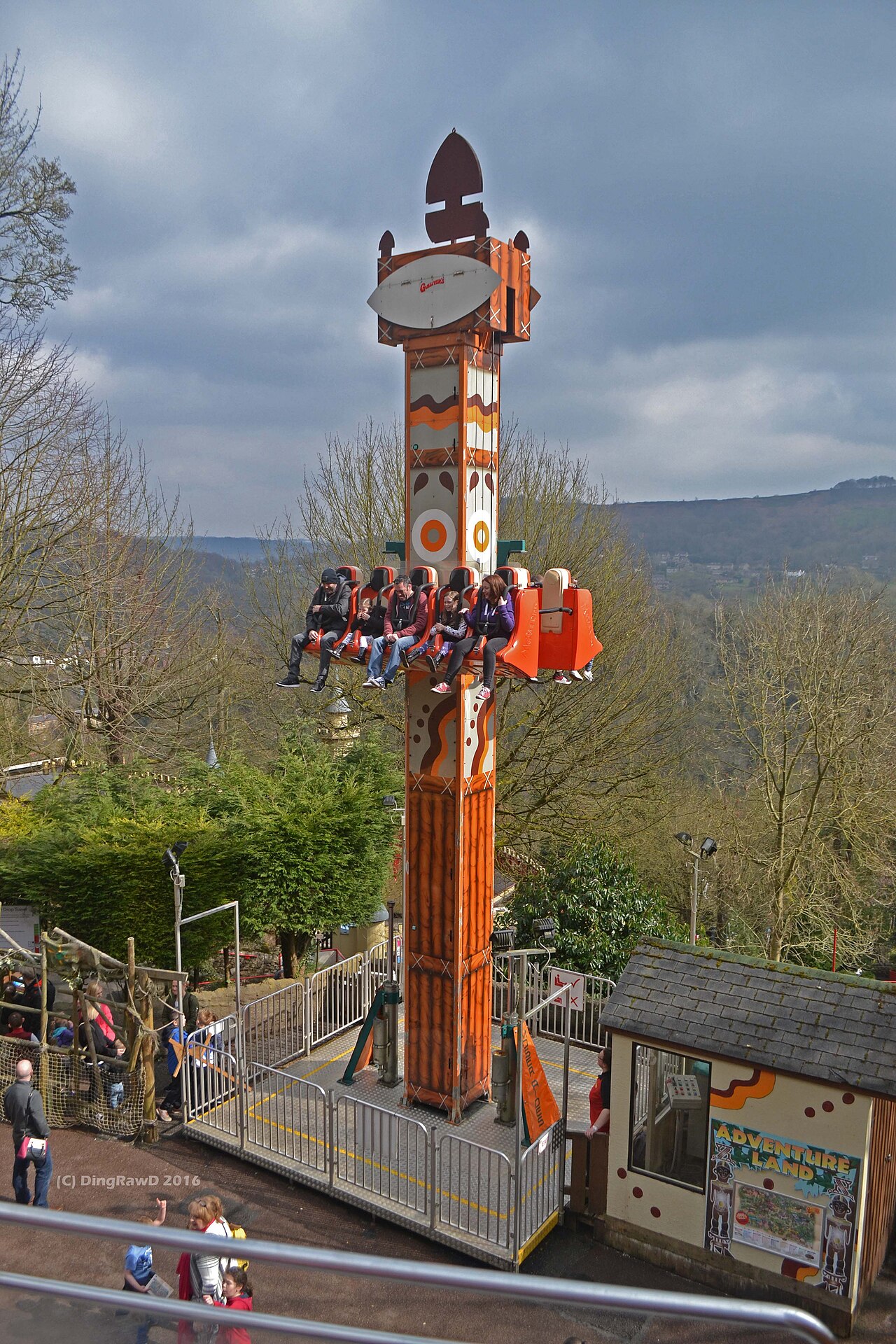 A tall vertical drop-tower ride with a row of seated riders carried partway up the central column.